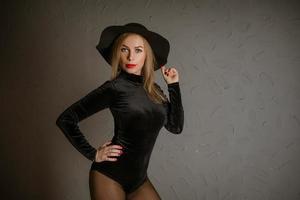 Portrait of a woman in a black bodysuit and hat posing against a wall photo