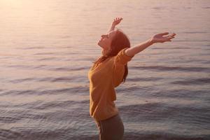 Young woman raises her hands to setting sun on shore lake, in yellow sweater photo