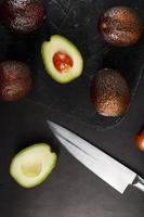 Whole and sliced avocados with a knife on a black textured black table, flat masonry. photo