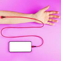 A smartphone with a large white screen is connected via a red USB cord to a vein in the hand on a Pink background. photo