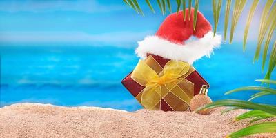 Red gift box with yellow bow and Santa Claus' hat on the beach behind sea and palm trees. Concept of Christmas, New year holiday in hot countries. Copy space photo