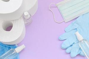 Set of important items for Covid-19 quarantine. Toilet paper, rubber disposable gloves with surgical face mask and hand sanitizer on lilac background photo