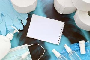 Set of items for Covid-19 quarantine. Empty notepad page and toilet paper, rubber gloves with surgical face mask and hand sanitizer with liquid soap bottle on human chest x-ray film photo