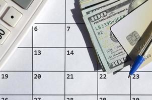 Pen and credit card on many hundred US dollar bills and calculator on calendar page close up photo