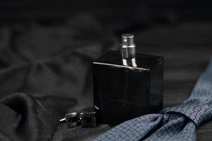 A bottle of mens cologne and cufflinks with blue tie lie on a black luxury fabric background on a wooden table. Mens accessories photo