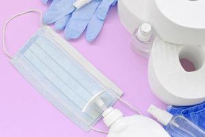 Set of important items for Covid-19 quarantine. Toilet paper, rubber disposable gloves with surgical face mask and hand sanitizer with liquid soap bottle on lilac background photo