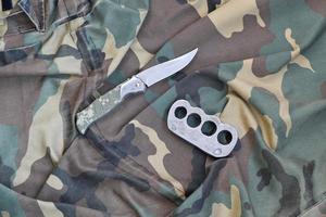 cuchillo y nudillos de latón sobre un fondo de ropa de camuflaje. herramientas para la autodefensa o ataques de delincuentes foto