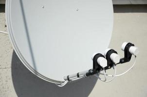 White satellite dish with three converters mounted on residental building rooftop concrete wall. Satellite television photo