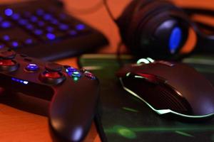 Modern gamepad and gaming mouse lies with keyboard and headphones on table in dark playroom scene. Cooperative teamplay concept photo