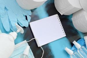 Set of items for Covid-19 quarantine. Empty notepad page and toilet paper, rubber gloves with surgical face mask and hand sanitizer with liquid soap bottle on human chest x-ray film photo