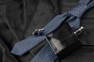 A bottle of mens cologne and cufflinks with blue tie lie on a black luxury fabric background on a wooden table. Mens accessories photo