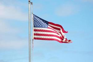 An old American flag on the tip of the flag with a torn in the wind on the flagpole against a blue sky background. photo