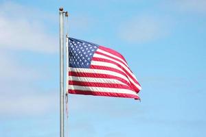 An old American flag on the tip of the flag with a torn in the wind on the flagpole against a blue sky background. photo