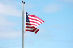 An old American flag on the tip of the flag with a torn in the wind on the flagpole against a blue sky background. photo