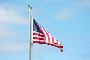 An old American flag on the tip of the flag with a torn in the wind on the flagpole against a blue sky background. photo