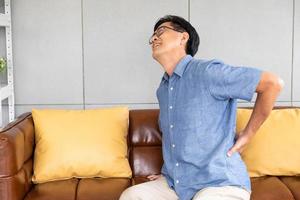 Indoor shot of senior Asian men sit on the sofa couch and touch his back and suffering from back pain, aching muscles, bones or lumbago. Elderly health problem concept. photo