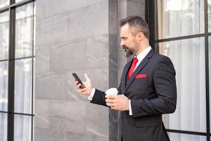 Selective focus at face. Adult Caucasian business man with beard, dress in formal suit standing in central business district. While drink morning coffee and look serious at his mobile smartphone. photo