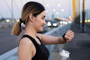 Selective focus at face of young beautiful Asian women using smart watch to track activity and listen music while warm up before exercise or running at the urban city view. Fit and healthy lifestyle. photo