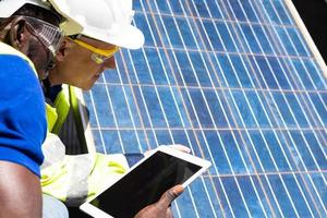 Selective focus at digital tablet. African and Caucasian engineer inspect electrical solar panel and record information into mobile tablet. Alternative energy and industrial concept. Outdoor shot. photo