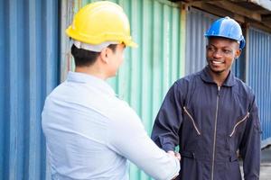 el enfoque selectivo en los trabajadores africanos se dan la mano con el supervisor de carga caucásico mientras trabajan en el sitio con casco de seguridad en el muelle logístico de importación o exportación de contenedores. trabajo en equipo industrial. foto