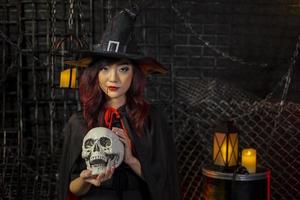 Chica asiática en traje de bruja sosteniendo el cráneo para el concepto de truco o trato de la fiesta de Halloween con fondo negro oscuro con espacio de copia foto