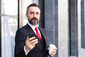 Selective focus at face. Adult Caucasian business man with beard, dress in formal suit standing in central business district. While drink morning coffee and look serious at his mobile smartphone. photo