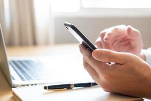 Selective focus at Smartphone. Men hand holding mobile phone while using finger to touch device screen. With blurred computer laptop and paper notebook with pen on working desk. Wireless technology. photo