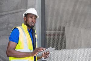 Selective focus at face of Black African foreman at building construction site, wearing protective hat and safety equipment while using digital tablet to record information. Civil engineer working. photo