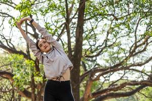 jóvenes y hermosas mujeres asiáticas que usan un reloj inteligente para rastrear la actividad y escuchar música desde un teléfono inteligente mientras se calientan y se estiran antes de hacer ejercicio de yoga con árboles naturales y de fondo. foto