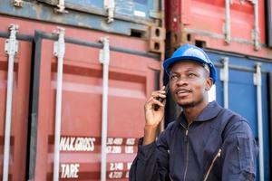 Selective focus at face of Black African man worker wearing safety equipment, talking on smartphone while check and inspection the condition of container. Import and export, logistic freight worker. photo