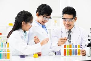 Young Asian kids wearing white scientist gown and do chemical test tube while study, learning in science classroom with teacher. Young children education concept by experiment, fun and enjoy classroom photo