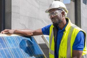 una foto al aire libre de un ingeniero africano negro inspecciona un panel solar eléctrico con casco, anteojos protectores y equipo de seguridad con una sonrisa en la cara. energía alternativa y concepto de trabajo industrial.