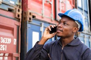 Selective focus at face of Black African man worker wearing safety equipment, talking on smartphone while check and inspection the condition of container. Import and export, logistic freight worker. photo