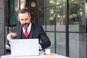 toma al aire libre de un hombre de negocios caucásico bebiendo café caliente por la mañana en la cafetería mientras usa una computadora portátil para trabajar y revisar su correo electrónico. rutina de trabajo con fondo de área de negocios urbanos. foto