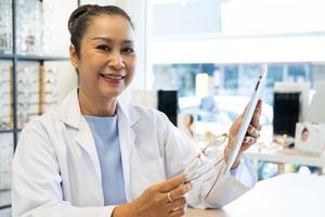 Asian women Doctor optometrist smiling while using tablet and recommend customer to choose glasses beautiful frame trial inside of optical shop, store. Eye sight for patient, healthcare concept. photo