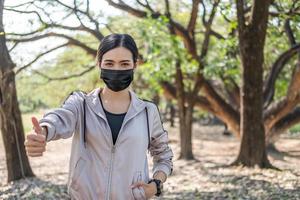 jóvenes y hermosas mujeres asiáticas que usan mascarilla quirúrgica y pulgares hacia arriba mientras usan un reloj inteligente para rastrear la actividad antes de hacer ejercicio o correr en el parque por la mañana. nuevo estilo de vida normal. foto