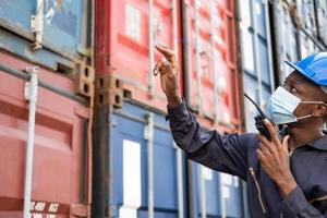 Selective focus at face of Black African man worker wearing surgical face mask and  safety equipment while check and inspection and using radio. New normal industrial container worker concept. photo