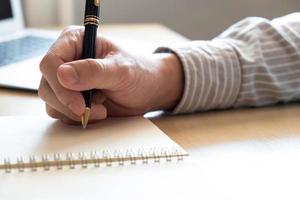 enfoque selectivo en la mano humana usando un bolígrafo para escribir una nota breve en papel en la mesa de trabajo. con computadora portátil borrosa en el fondo. lista para planificar u organizar la actividad durante los días laborables. foto