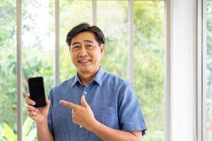 Asian elder man holding smartphone mock up with empty screen to add logo of application while point at that device with smile. Active senior  and technology with blurred green nature background. photo