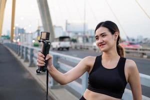 Selective focus at face of young beautiful Asian women using camera to online record and live stream while warm up before exercise or running at the urban city view. Fit and healthy lifestyle. photo