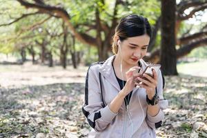 Selective focus at face of young beautiful Asian women using smart watch to track activity and listen music from smartphone while warm up before exercise yoga with natural and trees at background. photo