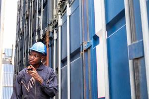 Selective focus at face of Black African logistic worker wearing safety equipment, check and inspection the condition of container while talking and communicate with his team by  radio device. photo