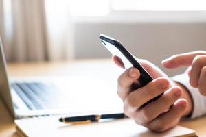 Selective focus at Smartphone. Men hand holding mobile phone while using finger to touch device screen. With blurred computer laptop and paper notebook with pen on working desk. Wireless technology. photo