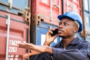 Selective focus at face of Black African man worker wearing safety equipment, talking on smartphone while check and inspection the condition of container. Import and export, logistic freight worker. photo