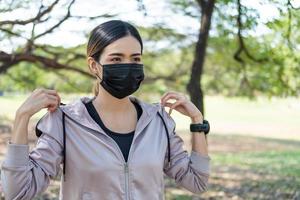 Selective focus at face of young beautiful Asian women wearing surgical face mask to prevent Covid-19 while warm up before exercise or running at the park in the morning. New normal lifestyle concept. photo