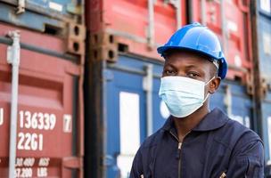 Selective focus at face of Black African man worker wearing surgical face mask and  safety equipment while check and inspection the condition of container. New normal industrial worker concept. photo