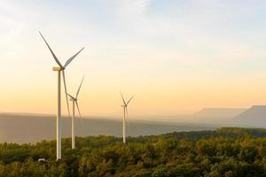 Landscape sun set shot of Wind turbines farm on the green grass field with clear blue sky and mountain at the background. Alternative green energy which generate electricity from natural wind power. photo