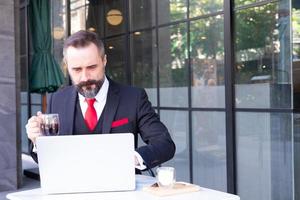toma al aire libre de un hombre de negocios caucásico bebiendo café caliente por la mañana en la cafetería mientras usa una computadora portátil para trabajar y revisar su correo electrónico. rutina de trabajo con fondo de área de negocios urbanos.