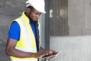 Selective focus at face of Black African foreman at building construction site, wearing protective hat and safety equipment while using digital tablet to record information. Civil engineer working. photo