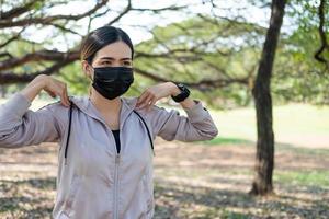 Selective focus at face of young beautiful Asian women wearing surgical face mask using smart watch to track activity before exercise or running at the park in the morning. New normal lifestyle. photo